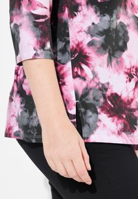 Květinový top s růžovými, černými a šedými odstíny, vyrobený z měkké tkaniny, s třičtvrtinovými rukávy a postranními rozparky, v kombinaci s černými kalhotami.