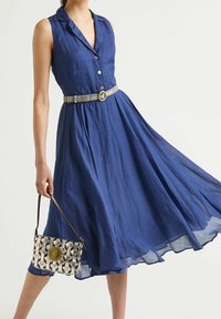 Ärmelloses, knielanges blaues Kleid mit Knopfleiste und tailliertem silbernen Gürtel; trägt eine gemusterte, metallische Handtasche mit einem runden Verschluss.