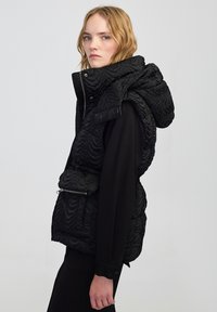 Czarna kamizelka puchowa o teksturowanej powierzchni, z oversize'owym kołnierzem i paskiem w talii. Zawiera kieszeń na zamek błyskawiczny oraz zapięcia na napy.