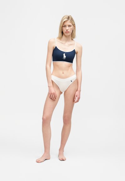 Νεαρή γυναίκα όρθια ξυπόλυτη, φορώντας μπλε ναυτικό κοντό τοπ με λευκό λογότυπο και λευκά σλιπ με ψηλή κοπή και μικρό μπλε λογότυπο σε απλό φόντο.