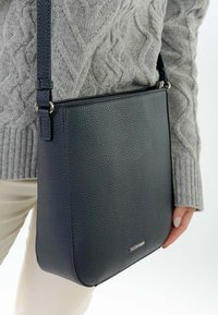 Person trägt grauen Strickpullover mit Zopfmuster und cremefarbene Hose und hält eine schwarze Schultertasche aus strukturiertem Leder mit silberner Logoschild.