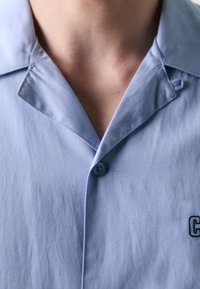 Vaaleansininen lyhythihainen paita, jossa on klassinen kaulus, napillinen etu ja pieni kirjailtu yksityiskohta vasemmalla puolella.