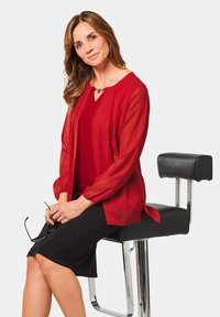 Rode chiffon blouse met lange mouwen over een rode top, gecombineerd met een zwarte rok tot de knie. Zittend op een zwarte stoel met chroomaccenten.