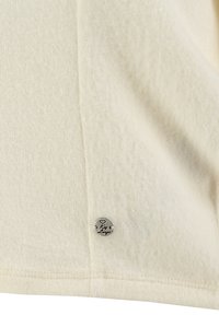 Tissu couleur crème avec couture visible et petite étiquette ronde en argent gravée de « Love » et d'un symbole de cœur.