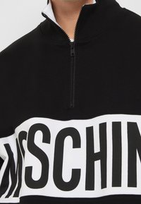 Černá zipová mikina s vysokým límcem, se širokým bílým pruhem přes hruď s černým textem. Hladký, elastický materiál.