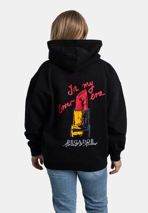 Person wearing black hoodie with red lipstick graphic and text "In my lover era" on back, paired with light blue jeans.
