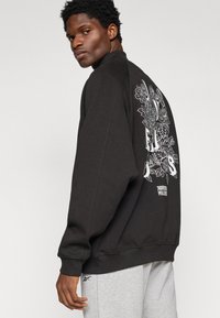 Felpa nera con grafica floreale bianca sul retro, vestibilità comoda, polsini e orlo a costine, abbinata a pantaloni della tuta grigi chiari.