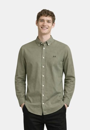 Giovane uomo sorridente, indossa una camicia color oliva chiaro con bottoni e un piccolo logo nero sul petto, mani nelle tasche, sfondo semplice.