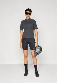 Kortermet, koksgrå sykkeltrøye, svarte polstrede shorts, svarte sokker og sykkelsko; iført solbriller, holder en grå hjelm.