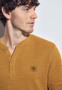 Mladý muž s krátkými hnědými vlasy, oblečený v hořčicově žlutém tričku s knoflíky a malou čtvercovou nášivkou na hrudi.