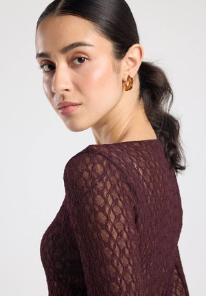 Bordeauxrotes Spitzenoberteil mit langen Ärmeln, versehen mit einem strukturierten Muster, rundem Ausschnitt und transparentem Detail. Kombiniert mit einem braunen Akzent-Ohrring.