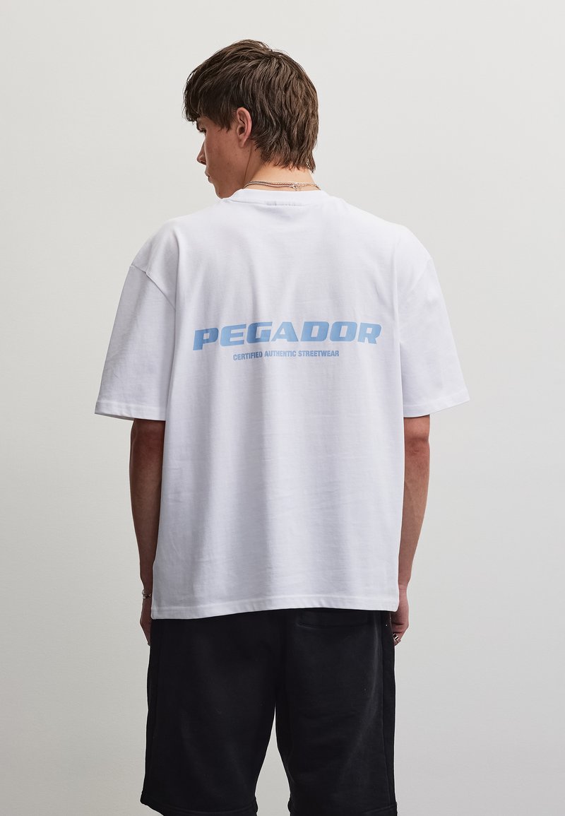 Νέος άνδρας με λευκό φαρδύ T-shirt με μπλε λογότυπο "PEGADOR" και μαύρα σορτς, γυρισμένος με την πλάτη σε ένα απλό φωτεινό φόντο.