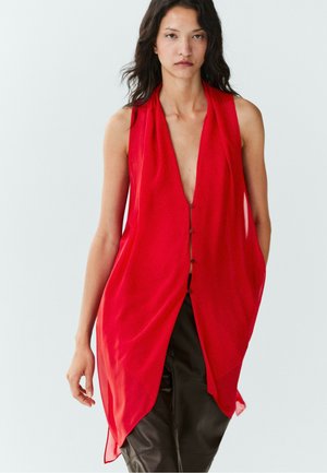 Femme aux longs cheveux foncés portant un top rouge sans manches avec un profond décolleté en V et un pantalon noir sur un fond clair uni.