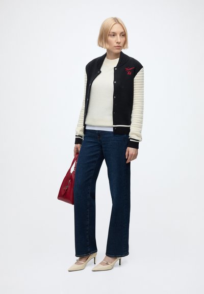 Femme aux cheveux blonds courts portant une veste universitaire noire et blanche, un haut blanc, un jean bleu, des talons beiges et tenant un sac à main rouge.