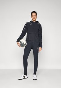 Mørk atletisk jakke med hvite detaljer og en halv glidelås-kollaret, kombinert med tettsittende bukser og hvite sportssko, holder en svart og hvit fotball.