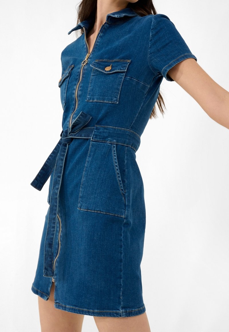 Femme portant une robe en jean bleue à manches courtes avec fermeture éclair à l'avant, poches à rabat sur la poitrine, poches latérales et ceinture nouée à la taille.