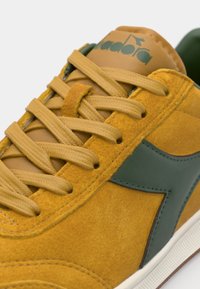 Baskets en daim jaune avec des accents en cuir vert, lacets plats et languette rembourrée avec détails de logo. Surface texturée avec des éléments contrastants.