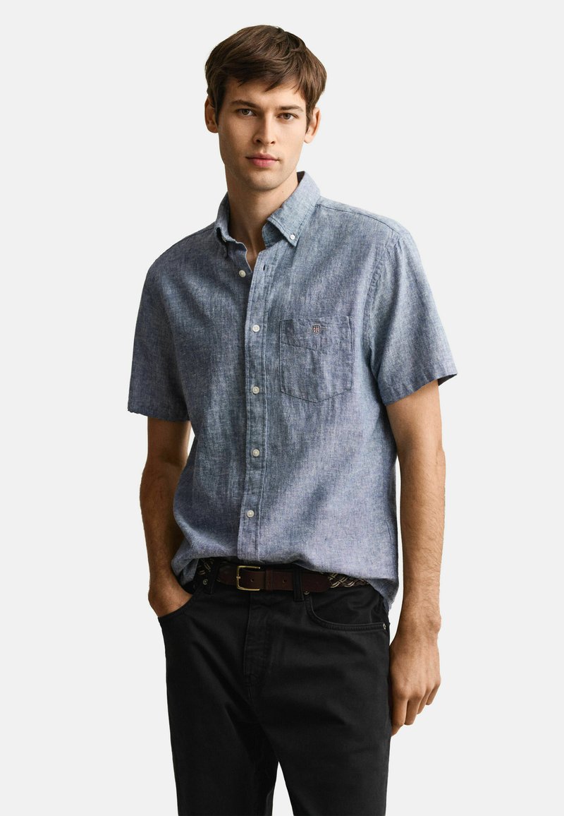 Giovane uomo che indossa una camicia blu a maniche corte con bottoni e taschino sul petto, pantaloni neri e cintura marrone, in piedi con una mano in tasca.