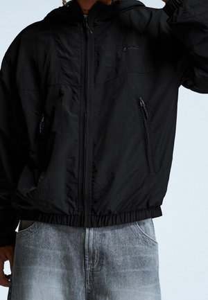 Personne portant une veste coupe-vent noire à fermeture éclair avec poches latérales zippées et un jean en denim bleu clair.