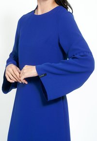 Rochie albastră vibrantă, cu mâneci lungi și decolteu rotund. Prezintă mâneci volan și o etichetă de marcă vizibilă la manșetă. Textură de țesătură netedă.