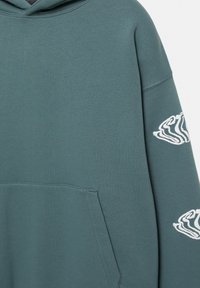 Sweat à capuche vert sarcelle avec impression textuelle blanche déformée sur la manche et une poche kangourou à l'avant.