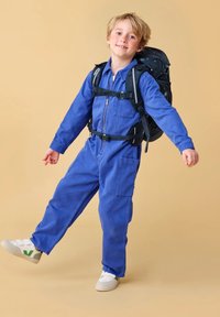 Mono de mezclilla azul con cremallera en la parte delantera, bolsillo en el pecho y grandes bolsillos laterales. Combinado con una mochila negra con un patrón de estrellas y zapatillas deportivas blancas.