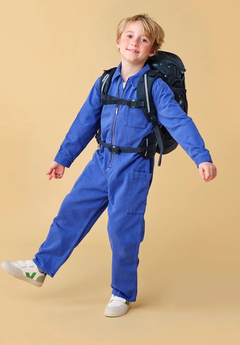 Mono de mezclilla azul con cremallera en la parte delantera, bolsillo en el pecho y grandes bolsillos laterales. Combinado con una mochila negra con un patrón de estrellas y zapatillas deportivas blancas.