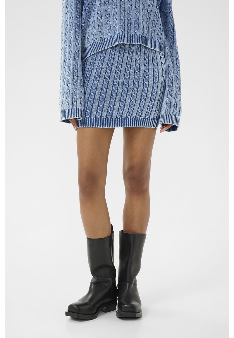 Femme portant un pull à torsades bleu et une mini-jupe assortie avec des bottes en cuir noires mi-mollets, debout sur un fond blanc.