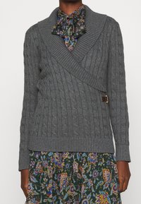 Tmavě pletá osoba nosící šedý svetr s pleteným vzorem přes zelené květinové šaty s kravatovou výstřih. G