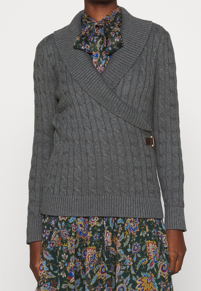 Tmavě pletá osoba nosící šedý svetr s pleteným vzorem přes zelené květinové šaty s kravatovou výstřih. G