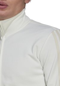 Biała sportowa kurtka na zamek z wysokim kołnierzem, prążkowaną fakturą i jasnobeżowymi paskami na ramionach. Gładkie wykończenie materiału.