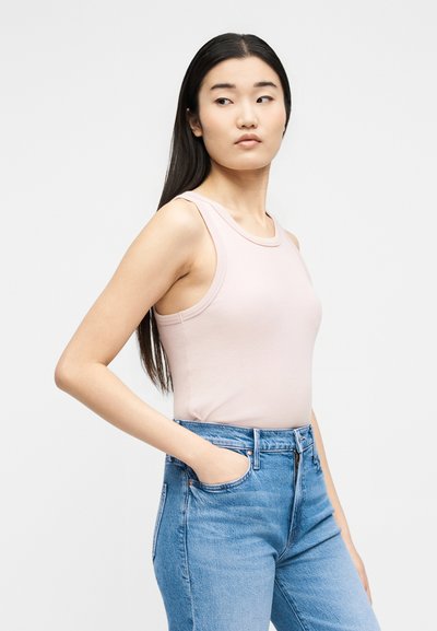 Νεαρή γυναίκα με μακριά σκούρα μαλλιά, φοράει αμάνικο ανοιχτό ροζ τοπ και μπλε τζιν, στέκεται με το ένα χέρι στην τσέπη κοιτάζοντας πλάγια.