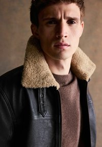 Jachetă din piele neagră cu un guler din blană bej. Purtați deasupra unui pulover maro, evidențiind texturi netede și o croială strânsă.