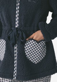 Giacca fuzzy blu scuro con una texture morbida, dotata di tasche a forma di cuore con motivo a scacchi e un elegante nodo di chiusura coordinato sul fronte.
