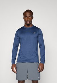 Mornarica dugih rukava sportska majica od prozračnog materijala s suptilnim uzorkom, u kombinaciji s svijetlosivim šortsevima, s oznakom New Balance.