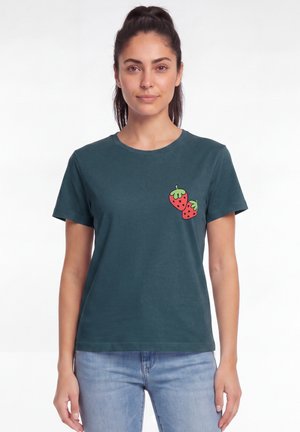 Frau mit dunkel zurückgebundenem Haar trägt ein dunkelgrünes T-Shirt mit zwei kleinen roten Erdbeeren auf der oberen linken Brust, kombiniert mit hellblauen Jeans.