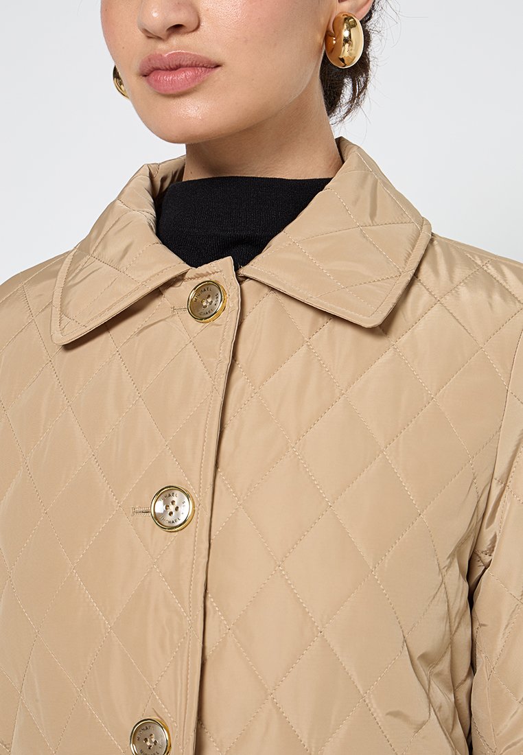 Giacca trapuntata beige con ampio colletto, dotata di bottoni dorati e una texture liscia e lucida. Indossata sopra una maglietta nera.