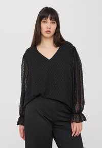 Blusa nera con scollatura a V, caratterizzata da motivi a pois texture e maniche lunghe trasparenti. Il design presenta polsini arricciati e una vestibilità comoda.