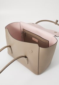 Sac fourre-tout en cuir beige avec deux poignées, ouverture sur le dessus, et un intérieur rose. Comprend une pochette zippée avec une étiquette de marque. Texture lisse, design minimaliste.