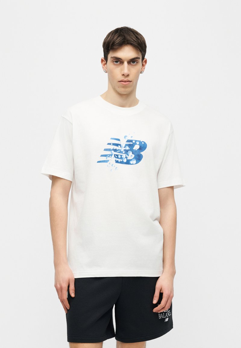 Jeune homme portant un t-shirt blanc avec un logo "NB" éclaboussé de bleu et un short noir avec un texte blanc sur une jambe, debout devant un fond uni.