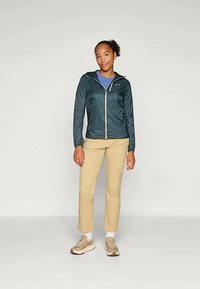 Lahka teal jakna s sprednjim zadrgo in kontrastnimi podrobnostmi zadrge, v kombinaciji s bež hlačami in nevtralnimi supergami.