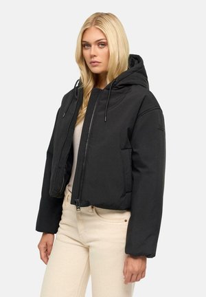 Blonde Frau mit schwarzer, gesteppter Kapuzenjacke mit Reißverschluss und beigen Hosen, die vor einem einfarbigen hellen Hintergrund steht.