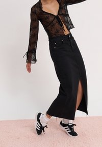Personne portant un haut transparent noir à fleurs à manches longues, une jupe midi en denim noir taille haute avec une fente à l'avant, des chaussettes blanches et des baskets Adidas noires.