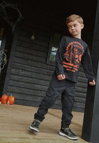 Svart långärmad skjorta med orange grafiskt tryck, kombinerad med svarta cargobyxor och gröna sneakers, placerad på en träveranda med pumpor.