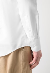 Weißes, strukturiertes Langarmhemd mit Knopfmanschette und abgerundetem Saum, kombiniert mit hellbraunen Hosen.