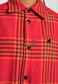 Rød og orangeternet flannel skjorte med sorte knapper, spidse krave og en brystlomme med et logotag. Blød tekstur.