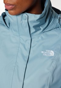 Gros plan d'une personne portant une veste The North Face bleu clair avec un col montant et un rabat avant boutonné.