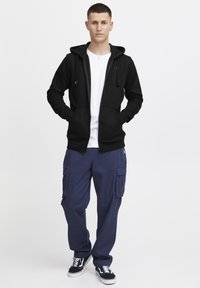 Haut à capuche noir zippé avec des cordons, porté sur un t-shirt blanc, associé à un pantalon cargo bleu marine et des baskets noires avec des accents blancs.