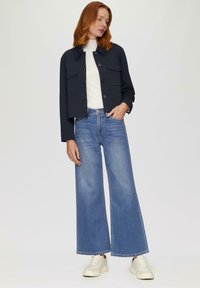 Vrouw met rood haar in een zwarte cropped jacket, witte top, wijdvallende blauwe jeans en witte sneakers, staande tegen een effen achtergrond.
