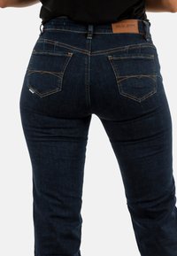 Jeans de corte slim de color azul oscuro, fabricados en denim, con bolsillos traseros con parches y costuras amarillas, y una etiqueta de cuero con marca en la cintura.